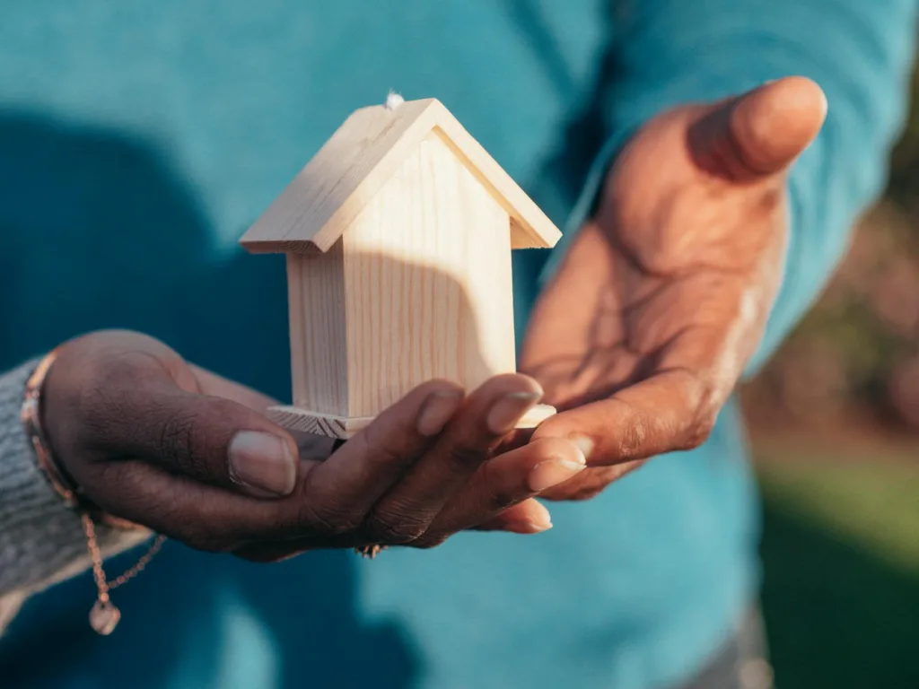 Close-up of hands holding a small wooden house model outdoors, symbolizing real estate investment.